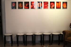 Ściana galerii oświetlona ciepłym światłem, na której w górnej części wisi poziomy rząd dziewięciu oprawionych fotografii. Zdjęcia przedstawiają abstrakcyjne, organiczne formy w intensywnych barwach czerwieni, pomarańczu i czerni, często przypominające przekroje owoców. Poniżej, na podłodze z beżowych płytek, stoi siedem czarnych taboretów z białymi siedziskami z czarnymi napisami, ustawionych w równym rzędzie. Po prawej stronie kadru widoczny jest fragment rzeźbionego fotela z tapicerką w musztardowym kolorze