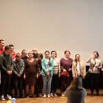 Grupa kilkunastu osób w różnym wieku, od dzieci po osoby starsze, stoi na scenie w rzędzie, z szerokim pustym ekranem w tle. Na pierwszym planie tyłem do widza stoi kobieta, robi zdjęcie lub filmuje