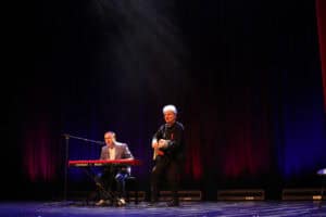Występ muzyczny Rafała Ugarenko: młody mężczyzna w okularach i jasnej marynarce siedzi po lewej stronie przy czerwonym keyboardzie (Nord Stage 3), gra na instrumencie i śpiewa do mikrofonu. W centrum sceny stoi ubrany na czarno Michał Stanowski, gra na gitarze akustycznej. W tle ciemna kurtyna oświetlona czerwono-niebieskimi światłami