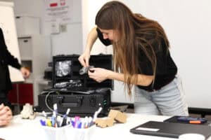 Kobieta z długimi włosami (Justyna Kazek-Góra) pochyla się nad otwartą czarną walizką ze sprzętem, wyjmuje małe, owinięte w folię elementy. Scena ilustruje przygotowania techniczne w trakcie szkolenia