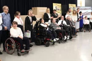 Szerokie ujęcie grupy osób na scenie. W pierwszym rzędzie siedzi sześć osób na wózkach inwalidzkich, a za nimi stoją ich opiekunowie. Tło tworzą stosy tekturowych pudeł z napisami, takimi jak „Konwencja" i „Pedagogika", oraz banery z chronologią wydarzeń historycznych stowarzyszenia. Po prawej widoczny jest fragment ekranu z napisem „WOLONTARIAT I PRAKTYKI STUDENCKIE"