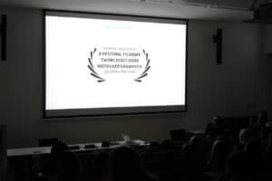 Pogrążona w ciemności sali wykładowa. Na dużym, jasnym ekranie wyświetlony jest napis: „Nagroda Publiczności X Festiwal Filmowy Twórczości Osób Niepełnosprawnych 2023 Biała Podlaska”. Na pierwszym planie widać zarys kilku rzędów publiczności