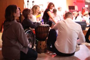 Zbliżenie na dwoje dorosłych i chłopca grającego na bębnie podczas koncertu. Chłopiec siedzi na wózku. Obok niego, na podłodze, siedzi dorosły mężczyzna w białej koszuli, tyłem do obiektywu oraz kobieta patrząca w prawą stronę. W tle widoczni są inni uczestnicy i instrumenty