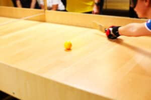 Zbliżone ujęcie drewnianego stołu do gry w showdown. W centrum leży żółta piłka. W prawym górnym rogu widać fragment ramienia i dłoni zawodniczki w czerwono-czarnej rękawicy, która trzyma rakietkę i zamierza uderzyć piłkę. Tło jest rozmyte, widoczne są fragmenty osób