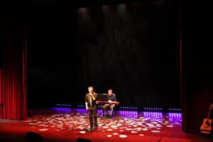 Występ Grzegorza Czapli. Na środku stoi młody mężczyzna w czarnej koszuli, śpiewa do mikrofonu. W tle, na podwyższeniu, siedzi muzyk grający na czerwonym keyboardzie. Scena teatralna oświetlona jest fioletowo-czerwonym światłem, podłogę sceny pokrywa wzór jasnych, okrągłych plam światła