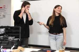 Demonstrowanie sprzętu do tłumaczenia. Młody mężczyzna w czarnej bluzie trzyma przy uchu słuchawkę nauszną, testując zestaw. Kobieta trzyma w dłoni mały odbiornik/nadajnik, uśmiecha się. Obydwoje stoją obok otwartej walizki z pętlą indukcyjną