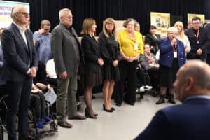 Grupa osób w strojach formalnych stojąca w półkolu na scenie. Po prawej, kobieta (Maria Król) w niebieskim żakiecie przemawia do mikrofonu, patrząc na publiczność znajdującą się poza kadrem. W tle widoczne są czarne zasłony, a także banery z historią stowarzyszenia