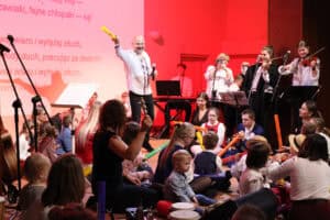 Mężczyzna w białej koszuli (Marek Fedor) stoi centralnie na scenie, trzyma mikrofon i unosi dłoń w geście dyrygowania. Na scenie siedzi duża grupa dzieci (niektóre w strojach ludowych i kapeluszach) oraz dorosłych, wspólnie biorą udział w występie, korzystając z instrumentów perkusyjnych. W prawym rogu sceny widoczny jest zespół składający się z dwóch skrzypaczek i kobiety robiącej zdjęcie telefonem. Na ekranie w tle widoczny jest fragment tekstu piosenki