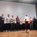 Ceremonia wręczenia upominków. Na scenie, na drewnianej podłodze przed dużym, białym ekranem, stoi ośmiu mężczyzn; po prawej stronie mężczyzna w białej koszuli i bordowych spodniach odbiera nagrodę, obok niego stoi chłopiec trzymający kolorową kartkę z napisem „DYPLOM"