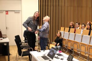 Sala wykładowa. Dwóch prelegentów stoi na pierwszym planie: mężczyzna z długimi włosami (Maciej Książko) i mężczyzna w koszuli w kratę (Piotr Kękuś), patrzą w górę z uśmiechem lub zaskoczeniem. Pomiędzy nimi widać zarys osoby siedzącej na wózku. Na pierwszym planie znajduje się białe biurko z otwartym laptopem. Po prawej widoczne są rzędy krzeseł, w których siedzi kilka kobiet, a na dole na podłodze, na wysokości pierwszego rzędu, siedzi młoda kobieta, patrząca w górę. Tło tworzy ściana z pionowymi drewnianymi panelami