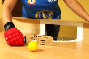 Bardzo zbliżone ujęcie piłki i rakietki do gry w showdown. Żółta piłka leży na drewnianym stole tuż obok drewnianej rakietki z widocznymi napisami i logo "Milo". Rakietka jest trzymana przez zawodniczkę w dużej, czerwono-czarnej rękawicy. Za rakietką i piłką widoczny jest otwór bramkowy i fragment siatki. Zawodniczka ma na sobie niebieską koszulkę