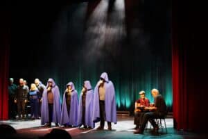 Scena podczas występu WTZ „Misericordia”: po lewej stronie w tle stoi grupa chórzystów w czarnych strojach, w centrum cztery osoby w długich fioletowych pelerynach z kapturami śpiewają do mikrofonów, a po prawej dwie osoby siedzą i akompaniują na gitarach