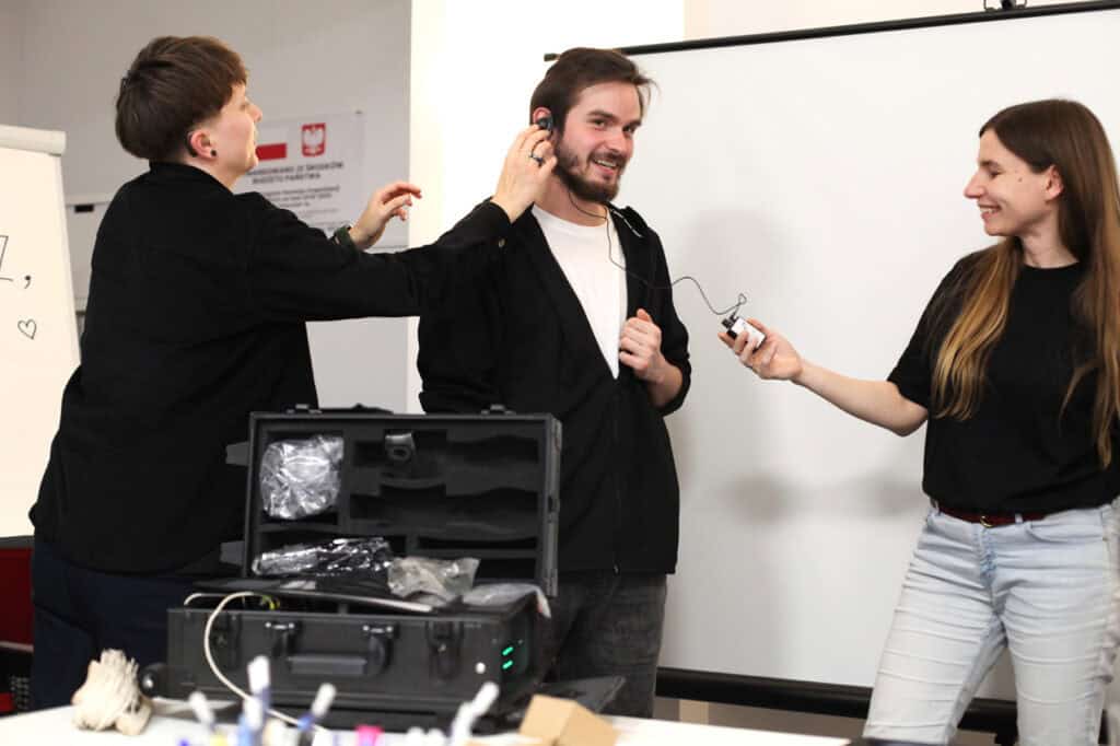 Testowanie sprzętu w trakcie szkolenia. Uśmiechnięty mężczyzna ma na sobie słuchawkę nauszną. Prowadząca (po lewej) pomaga mu w regulacji, a kobieta (po prawej) trzyma nadajnik, uśmiechając się do niego. W tle widać walizkę z otwartym sprzętem (pętla indukcyjna)