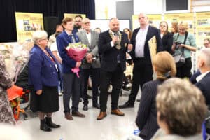 Ceremonia wręczenia wyróżnień. Przed publicznością stoi grupa osób w strojach formalnych. W centrum do mikrofonu przemawia mężczyzna w ciemnym garniturze. Po jego prawej stoi wysoki mężczyzna, trzyma oprawiony obraz lub dyplom. W tle widoczne są banery z historią stowarzyszenia. Na pierwszym planie widoczne są tyły głów publiczności