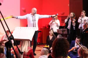 Mężczyzna w białej koszuli z haftem (Marek Fedor) stoi na scenie. Ma rozłożone ręce - w prawej trzyma żółty instrument w kształcie rurki, w lewej mikrofon. Wokół niego siedzą dorośli i dzieci. Po prawej stronie w tle widoczny jest zespół składający się z dwóch skrzypaczek i wokalistki. Na ekranie w tle widoczne są fragmenty tekstu piosenki