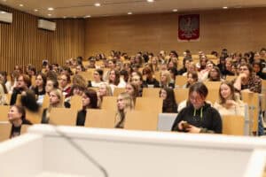 Szeroki kadr przedstawiający aulę wykładową wypełnioną studentami. Większość publiczności to młode kobiety, skupione na prelekcji. Siedzą w rzędach krzeseł z drewnianymi oparciami i blatami. Ściany i sufit są wykończone jasnym drewnem i panelami. Z tyłu sali, centralnie, na drewnianym panelu, wisi godło Polski. Na pierwszym planie widoczny jest fragment białego stołu lub pulpitu.