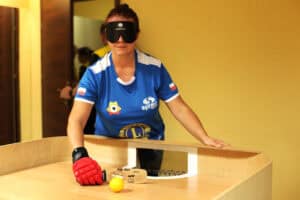 Zawodniczka (Elżbieta Mielczarek) grająca w showdown. Kobieta w niebieskiej koszulce sportowej z flagą Polski i czarnej opasce na oczach, stoi za drewnianym stołem do gry. Trzyma rakietkę w dłoni w dużej, czerwono-czarnej rękawicy tuż przy żółtej piłce, gotowa do uderzenia w kierunku bramki