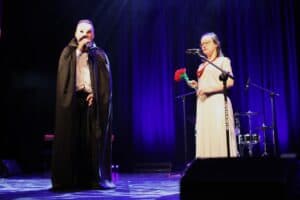 Występ Kingi Gańskiej i Łukasza Liska. Dwoje wykonawców śpiewa na scenie z niebieską kurtyną w tle. Mężczyzna po lewej stronie jest przebrany za upiora w masce, białej koszuli i czarnej pelerynie. Kobieta po prawej, w długiej, białej sukni i czerwonej ozdobie na szyi, trzyma w ręce bukiet sztucznych czerwonych róż i śpiewa do mikrofonu