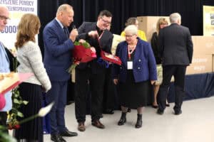Ceremonia wręczania wyróżnień. Od lewej stoi mężczyzna w granatowym garniturze. Trzyma mikrofon i duży bukiet czerwonych kwiatów. W centrum stoi kobieta (Maria Król) w niebieskim, tweedowym żakiecie, spogląda na otwarte pudełko trzymane przez stojącego obok mężczyznę (Kamil Kubasiński) w okularach i czarnym garniturze. W tle widoczne są stosy tekturowych pudeł z napisami m.in.: „ICF" i „Wsparcie Węgry"