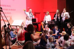 Aktywny moment na scenie oświetlonej na czerwono. Mężczyzna (Marek Fedor) w białej koszuli śpiewa do mikrofonu. Tuż obok niego stoi mały chłopiec w białej koszuli, patrzy w górę. Wokół nich siedzi duża grupa dzieci (w tym kilkoro w strojach galowych i ludowych) oraz dorosłych. W tle widoczny jest zespół składający się z dwóch skrzypaczek i wokalistki w białej bluzce. Na ekranie w tle widoczny jest tekst piosenki