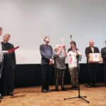 Osiem osób stoi na scenie podczas ceremonii wręczenia nagród: po lewej mężczyzna w koszuli i jeansach z mikrofonem i czerwoną teczką w dłoni; obok ksiądz w sutannie, a po prawej grupa laureatów i organizatorów, z dwoma kobietami trzymającymi statuetki i dyplom oraz mężczyzną z pudełkiem z czerwonym logo