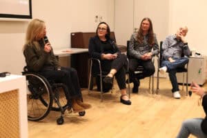 Panel dyskusyjny w sali wykładowej. Cztery osoby siedzą na krzesłach. Skrajnie po lewej, młoda kobieta na wózku (Angelika Chrapkiewicz-Gądek), ubrana w ciemnobrązowy sweter, trzyma mikrofon i przemawia. Obok niej siedzi kobieta w okularach i ciemnym żakiecie, wpatruje się w prelegentkę (Anna Bieganowska-Skóra). Obok z rękami skrzyżowanymi na kolanach siedzi mężczyzna z długimi włosami (Maciej Książko). Skrajnie po prawej, mężczyzna w koszuli w kratę siedzi z nogą założoną na nogę, zamyślony, z ręką na podbródku (Piotr Kękuś)