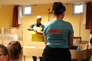 Ujęcie z tyłu na zawodniczkę w jasnoniebieskiej koszulce z napisami na plecach (w odbiciu lustrzanym), stojącą przy drewnianym stole do showdown. W tle, za stołem, widać mężczyznę w żółto-czarnej koszulce (sędzia) trzymającego mały przedmiot. W pomieszczeniu znajdują się dwa małe okna z bordowymi zasłonami po bokach