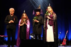 Cztery osoby stoją obok siebie na scenie po występie. Po lewej Michał Stanowski w czarnym stroju, z odznaczeniem przypiętym na piersi. Obok niego trzy kobiety/dziewczyny w czarodziejskich czapkach i przebraniach czarownic. Jedna z nich ma fioletową pelerynę, a inna czarną. Każda trzyma w dłoni czerwoną różę