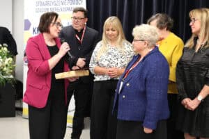 Grupa sześciu osób na scenie podczas uroczystości. Z lewej strony stoi kobieta (Anna Bieganowska-Skóra) w marynarce w kolorze fuksji i okularach, trzyma mikrofon i ozdobne pudełko. Zwraca się do kobiety (Maria Król) w niebieskim, tweedowym żakiecie. Pomiędzy nimi stoi kobieta o jasnych włosach w biało-czarnej bluzce. W tle widoczni są: mężczyzna w okularach (Kamil Kubasiński) i garniturze (częściowo zasłonięty), kobieta w żółtej bluzce oraz kobieta w czarnej, teksturowanej sukience. Po lewej stronie tła widoczny jest baner Stowarzyszenia Pomocy Dzieciom Niepełnosprawnym „Krok za Krokiem"