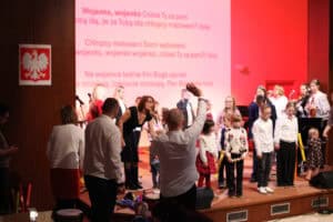 Mężczyzna w białej koszuli stoi tyłem do obiektywu, jest zwrócony w stronę sceny. Z uniesioną prawą ręką dyryguje lub zachęca do śpiewu dzieci i dorosłych, znajdujących się na scenie. Dzieci są ubrane w galowe stroje (białe koszule, ciemne spodnie/spódnice). Po lewej stronie wisi godło Polski. Na dużym ekranie w tle wyświetlany jest tekst piosenki "Wojenko, wojenko cóżeś Ty za pani?"