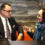 Mężczyzna (Jakub Semeniuk) w ciemnym garniturze i okularach, siedzi na pomarańczowym fotelu, rozmawia z kobietą w okularach (Anna Bieganowska-Skóra), w szalu i kurtce, która trzyma w dłoniach mały, czerwony przedmiot, prawdopodobnie dyktafon lub telefon komórkowy. W tle widoczne są marmurowe schody