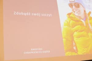 Zbliżenie na slajd prezentacji, utrzymany w jasnej, pomarańczowo-różowej kolorystyce. Po lewej stronie widoczny jest tekst: „Zdobądź swój szczyt", a na dole „ANGELIKA CHRAPKIEWICZ-GĄDEK". Po prawej stronie slajdu znajduje się jasne zdjęcie Angeliki Chrapkiewicz-Gądek ubranej w żółtą, puchową kurtkę, niebieską czapkę i okulary przeciwsłoneczne, uśmiechającej się w plenerze, prawdopodobnie w górach