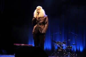 Mężczyzna na scenie w kostiumie fantasy, z długą, jasną (platynową) peruką i w ciemnej, skórzanej tunice z paskiem, mówi do mikrofonu. W tle widoczny zestaw perkusyjny i fragment keyboardu. Kurtyna oświetlona jest niebieskim światłem