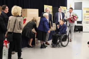 Mężczyzna (Robert Jagodziński) w niebieskiej marynarce siedzi na wózku, przemawia do mikrofonu na scenie. Kobieta (Maria Król) w niebieskim żakiecie pochyla się w jego kierunku. Wokół stoi grupa osób. Po prawej stronie dwaj mężczyźni w garniturach, z czego jeden trzyma dyplom i duży bukiet kwiatów. W tle widoczne są pudła i banery z historią stowarzyszenia. Po lewej stronie, tyłem do obiektywu, stoi kobieta w beżowej, wzorzystej marynarce i czarnych spodniach