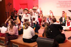 Zbliżenie na środkową część sceny oświetlonej na czerwono. W centrum uśmiechnięty mężczyzna w tradycyjnej białej koszuli z haftem, otoczony dwoma małymi chłopcami ubranymi w podobne, tradycyjne stroje ludowe i słomiane kapelusze. Wokół nich na podłodze sceny siedzi grupa dzieci i dorosłych biorących udział w występie. W tle widoczni są inni muzycy.