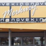 Zewnętrzny widok na wejście do kina z dużym, poziomym szyldem w kolorze żółtym i czarnym, na którym białymi, stylizowanymi literami widnieje nazwa "Merkury" oraz powtarzający się napis "Nove Kino"; po prawej stronie banera widoczny jest adres internetowy kinomerkury.pl