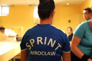 Ujęcie z tyłu na zawodniczkę w niebieskiej koszulce z białym napisem "SPRINT WROCŁAW" na plecach. Kobieta ma włosy upięte w kok. W tle, po lewej, widać zamazaną postać pochyloną nad stołem do gry w showdown