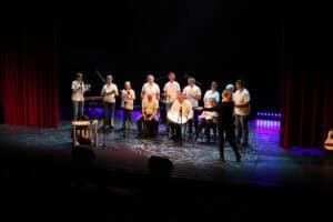 Szerokie ujęcie sceny, na której występuje grupa "Bębnoludy" z WTZ w Janowicy. Większość osób jest ubrana w białe koszulki i gra na nietypowych instrumentach, w tym na tubach perkusyjnych i białych bębnach obręczowych. W tle widoczny fioletowy pas świateł LED i czerwone kurtyny po bokach