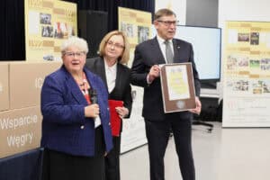 Trzy osoby stoją na scenie podczas uroczystości. Po lewej, kobieta (Maria Król) w niebieskim, tweedowym żakiecie i okularach przemawia do mikrofonu. Obok niej stoi kobieta (Małgorzata Romanko) w czarnym garniturze i okularach, trzyma czerwoną teczkę. Po prawej, mężczyzna w ciemnym garniturze i krawacie trzyma oprawiony w ramkę „MEDAL PAMIĄTKOWY WOJEWÓDZTWA LUBELSKIEGO”. W tle widoczne są stosy tekturowych pudeł z napisami m.in.: „ICF" i „Wsparcie Węgry" oraz pionowe banery z historią stowarzyszenia w ujęciu chronologicznym od 1990 roku