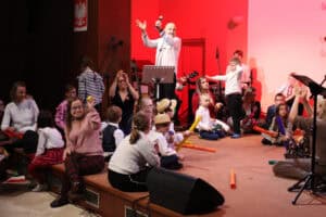 Mężczyzna w białej koszuli (Marek Fedor) stoi na scenie i macha rękami nad głową. Wokół niego, na scenie oświetlonej na czerwono, siedzi duża grupa dzieci i dorosłych (w tym kilka osób w strojach ludowych), bawiących się kolorowymi instrumentami. Na pierwszym planie, z prawej strony, kobieta siedzi tyłem i klaszcze, a z lewej inna kobieta uśmiecha się i trzyma żółtą piłeczkę. Po lewej stronie, w górnym rogu wisi godło Polski