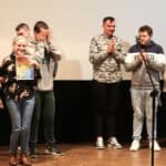 Widok na scenę. Od lewej dwie uśmiechnięte kobiety, z których jedna trzyma dyplom. Kolejno kilka osób klaszczących w dłonie. Z prawej ksiądz w sutannie (Paweł Zazuniak). Wszyscy stoją na jasnej, drewnianej podłodze, na tle białego ekranu