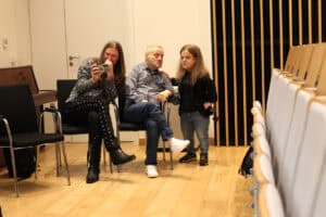 Trzy osoby podczas rozmowy na sali wykładowej. Po lewej stronie mężczyzna (Maciej Książko) z długimi włosami siedzi na krześle, trzyma telefon komórkowy (prawdopodobnie robi zdjęcie) i uśmiecha się. Kolejno, mężczyzna (Piotr Kękuś) w koszuli w kratę siedzi z założonymi nogami i rozmawia. Po prawej, młoda kobieta (Anna Dzieduszycka), ubrana w czarny żakiet i dżinsy, stoi obok niego, opierając rękę na jego ramieniu. Tło stanowi jasna, drewniana podłoga, rzędy krzeseł z prawej strony i ściana z pionowymi drewnianymi panelami