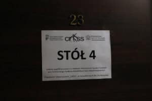 Tabliczka informacyjna "STÓŁ 4" przyczepiona do ciemnych, drewnianych drzwi oznaczonych złotym numerem "23". Na tabliczce widnieją logotypy Państwowego Funduszu Rehabilitacji Osób Niepełnosprawnych, Ministerstwa Sportu i Turystyki oraz Stowarzyszenia "CROSS". Poniżej znajduje się informacja o współfinansowaniu zadania i dane organizatora: Stowarzyszenie "CROSS" z Warszawy