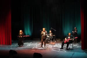 Na scenie występ zespołu "Siódme Niebo" z WTZ Caritas w Chełmie. W centrum stoi wokalista w czarnej kurtce, śpiewający do mikrofonu. W tle za nim siedzi perkusista a obok gitarzysta basowy z czerwoną gitarą. Po bokach dwie kobiety w długich, czarnych sukniach, z których jedna gra na gitarze. Tło stanowi ciemnoturkusowa kurtyna