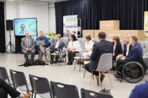 Panel dyskusyjny. Dziewięć osób (w tym jedna na wózku inwalidzkim) siedzi w półkolu na szarych sofach i fotelach, rozmawiając. Na podłodze, przed mężczyzną siedzącym po lewej stronie (Paweł Wdówik), leży czarny pies-przewodnik. W tle po lewej widoczny jest duży ekran z prezentacją przedstawiającą portret mężczyzny w okularach. Obok stoi pionowy baner Stowarzyszenia Pomocy Dzieciom Niepełnosprawnym. Z tyłu widać rząd tekturowych pudeł z napisami „Konwencja", „Rehabilitacja", „AAC" i „ICF"