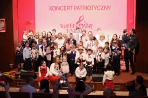 Fotografia grupowa zespołu, uczestników, dzieci i rodziców na scenie. Wszyscy stoją lub siedzą blisko siebie, uśmiechając się. W centralnym punkcie stoją dzieci w tradycyjnych strojach ludowych i kapeluszach. Na ekranie w tle widnieje napis „KONCERT PATRIOTYCZNY" i logo „The Fedor Family". Na pierwszym planie widoczna jest publiczność robiąca zdjęcia telefonami