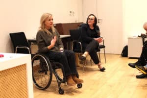 Od lewej młoda, blondwłosa kobieta (Angelika Chrapkiewicz-Gądek) w ciemnym swetrze i brązowych botkach siedzi na wózku i gestykuluje prawą dłonią. Obok niej siedzi kobieta w okularach i ciemnym żakiecie (Anna Bieganowska-Skóra), spogląda w prawo. Podłoga jest jasna, drewniana. W tle widoczne są biurka i ciemne pianino.