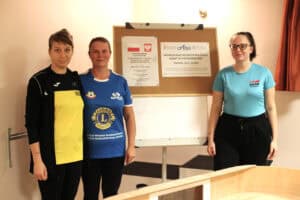 Trzy kobiety (Jagoda Grudzień, Elżbieta Mielczarek, Weronika Wall) pozują na tle tablicy informacyjnej. Z lewej, kobieta w czarno-żółtej bluzie. W środku, kobieta w niebieskiej koszulce sportowej z logo "Lions International". Po prawej, kobieta w okularach i jasnoniebieskiej koszulce z napisem "DSAN". Na tablicy, za nimi, widoczny jest plakat promujący "Indywidualne Mistrzostwa Polski Kobiet w Showdown 2025, z logotypami PFRON, CROSS i Ministerstwa Sportu i Turystyki. Na dole widoczna jest krawędź stołu do showdown