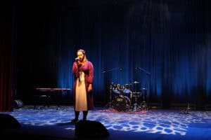 Występ Julii Majsterek. Kobieta stoi samotnie na scenie w świetle reflektorów, śpiewa do mikrofonu. Ubrana jest w długą, bordową sukienkę. W tle widoczny zestaw perkusyjny i keyboard. Scena jest oświetlona na niebiesko, a na podłodze widoczne są jasne, nieregularne plamy światła