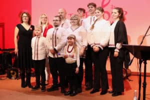 Zbliżenie na kluczowych członków zespołu i organizatorów, stojących razem na scenie. Grupa składa się z jedenastu osób (dzieci i dorosłych). Większość ubrana jest w białe koszule z czerwono-czarnym haftem, symbolizującym tradycyjne stroje ludowe. Dziewczynka w pierwszym rzędzie trzyma kolorową rurkę i ma na sobie czerwone korale. W tle ekran oświetlony jest na czerwono